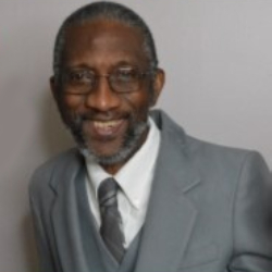 A man with short gray hair and a beard smiles at the camera. He is wearing glasses, a white shirt, a gray tie, and a gray suit jacket. The background is a plain, light-colored wall, emphasizing his composed demeanor as he advocates for water donation initiatives.