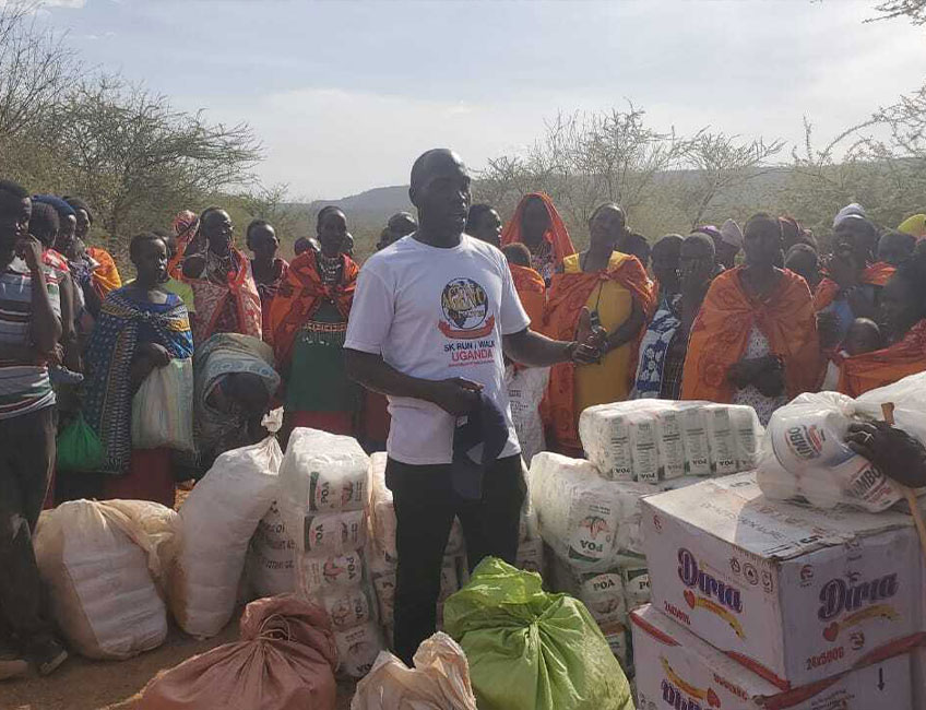 A man stands among numerous boxes and bags labeled “Dina,” surrounded by a group dressed in colorful clothing, some holding bags and other items. Addressing the crowd outdoors near sparse trees, he appears to lead a charity drive, encouraging them to donate water and supplies.