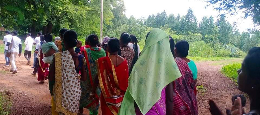 A group of people, mostly women wearing colorful sarees, walk down a dirt path surrounded by lush greenery. Some men in white shirts are walking ahead of them. The scene appears to be in a rural setting where the community thrives on charity and donations.