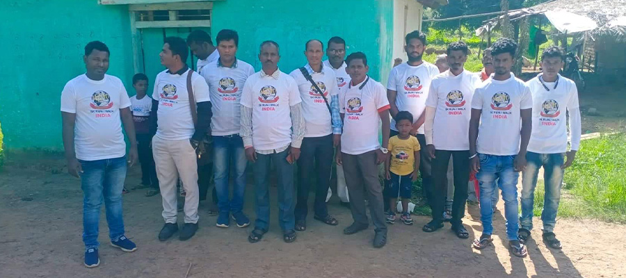 A group of men and a couple of children are standing outside in front of a turquoise building. The men, who likely participate in local charity efforts, are wearing white t-shirts with a printed design. Some are smiling at the camera while others have neutral expressions, adding to the rural setting's charm.